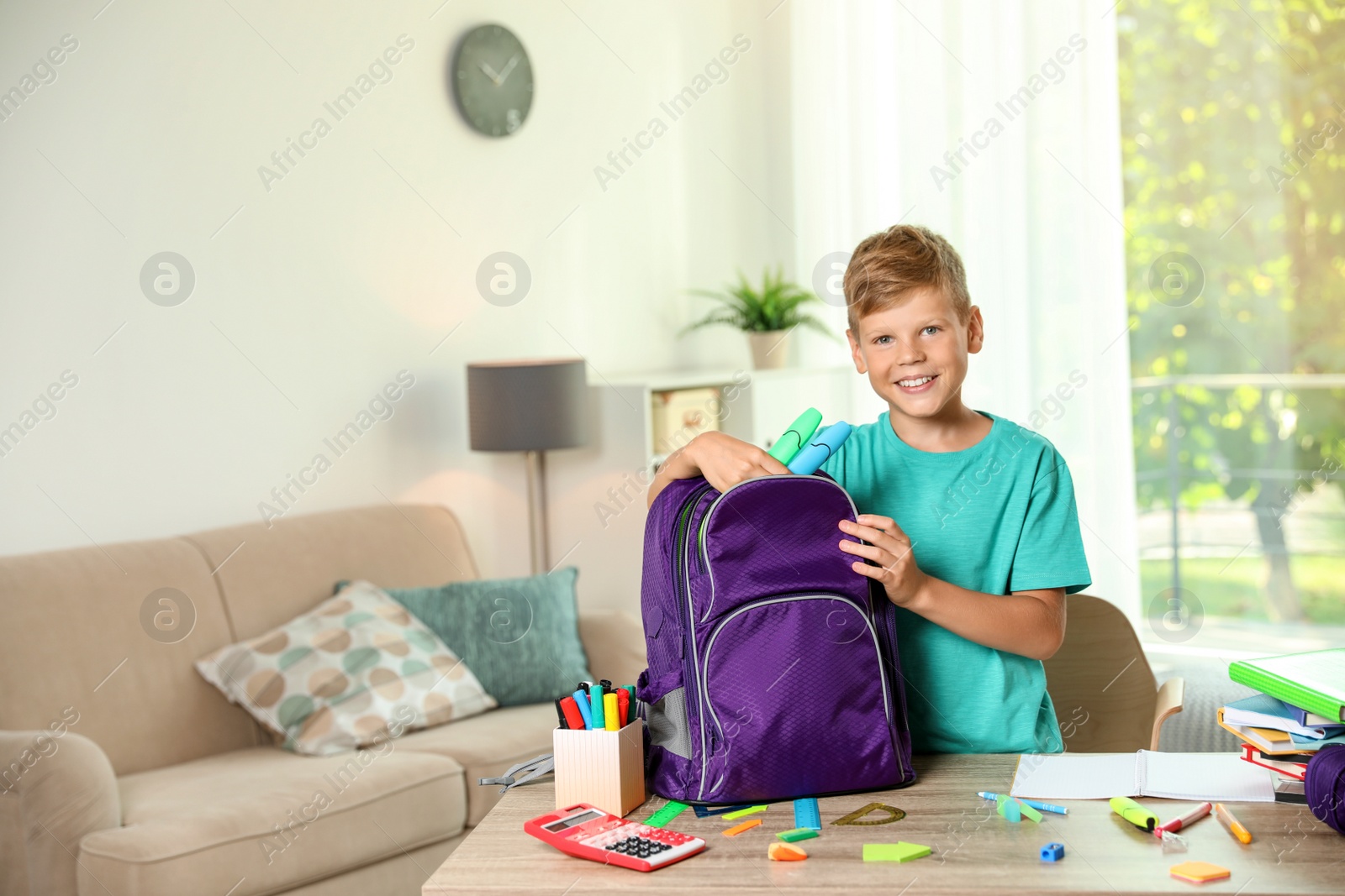 Photo of Cute boy putting school stationery into backpack at table indoors