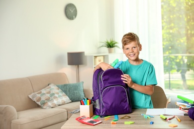Photo of Cute boy putting school stationery into backpack at table indoors