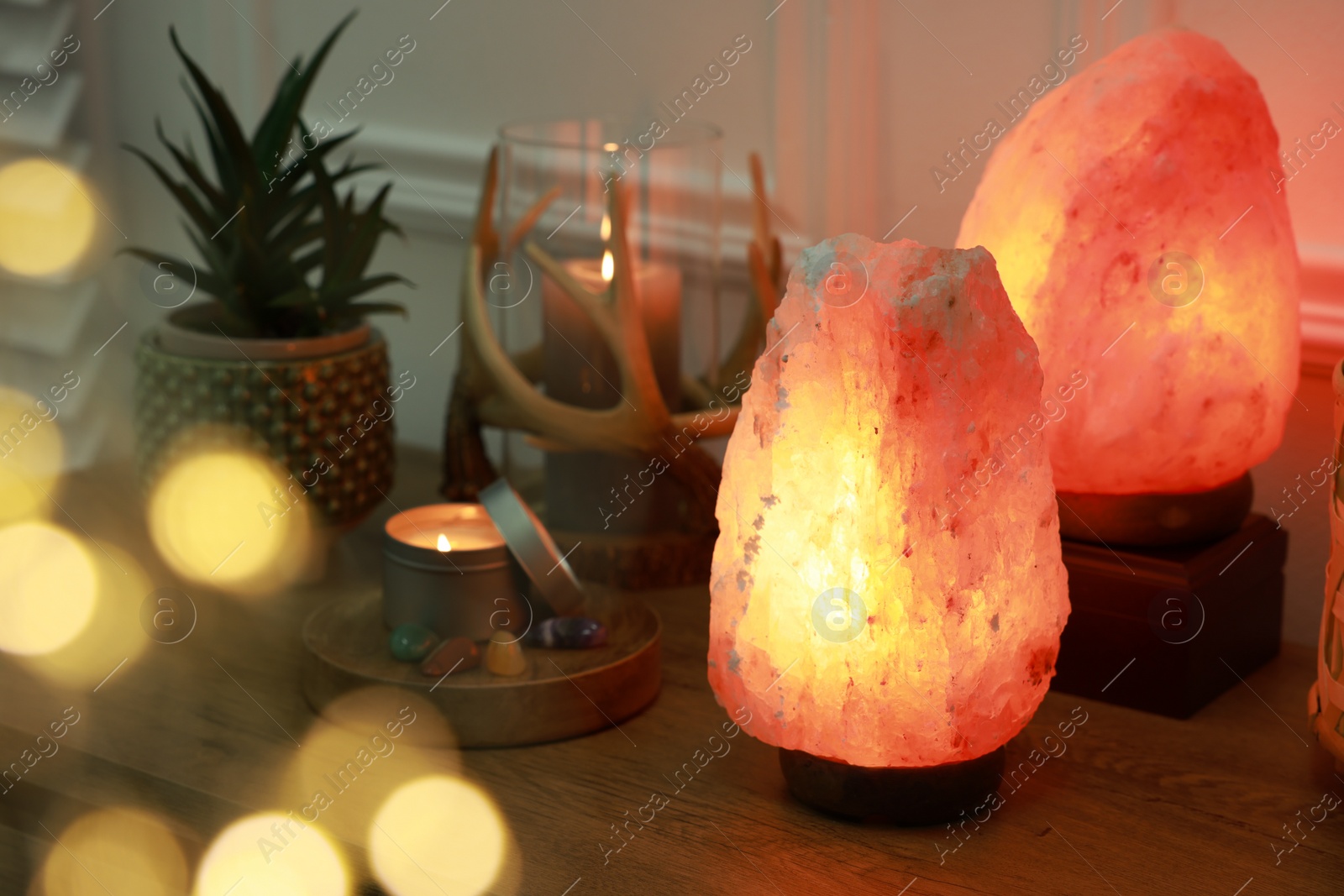 Photo of Himalayan salt lamps, candles, houseplant and gemstones on wooden table near white wall indoors. Bokeh effect