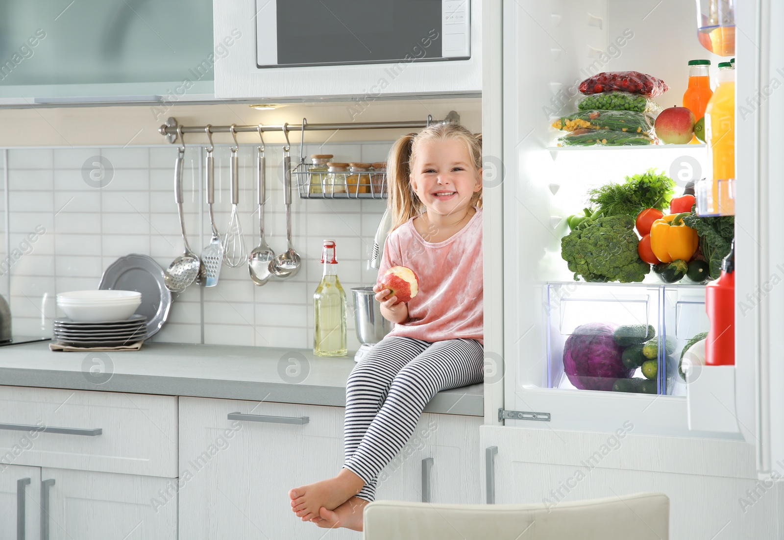 Photo of Cute little girl with apple sitting near open refrigerator in kitchen