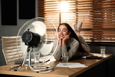 Woman enjoying air flow from fan while lying on table at workplace. Summer heat