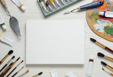 Photo of Blank canvas, tools, palette and paints on white wooden table, flat lay. Space for text