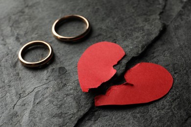 Photo of Halves of torn red paper heart and wedding rings on dark grey table. Broken heart