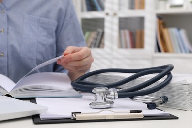 Photo of Woman with book, stethoscope and clipboard at white table indoors, closeup. Medical education