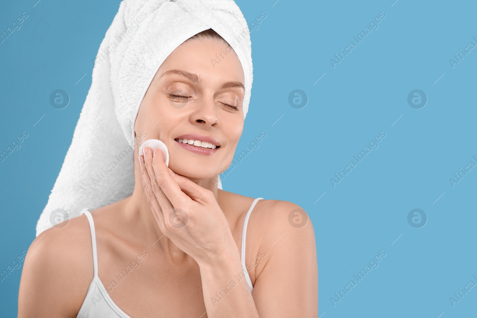 Photo of Beautiful woman in terry towel removing makeup with cotton pad on light blue background, space for text