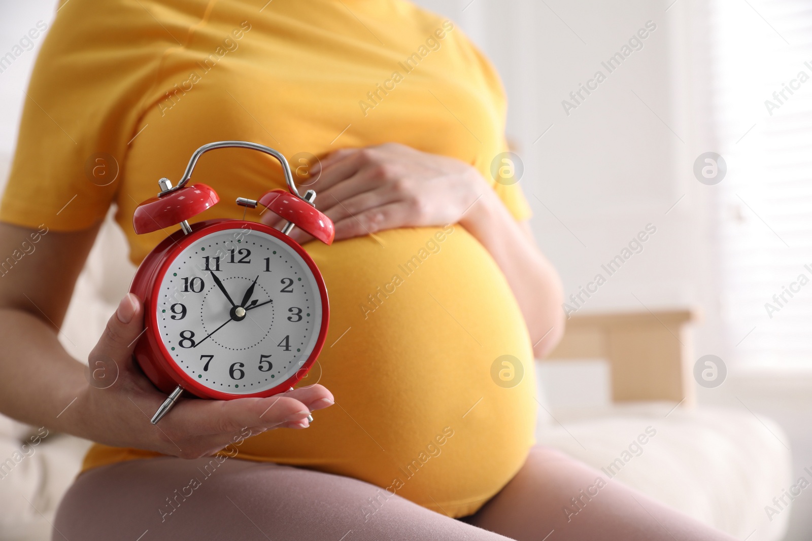 Photo of Young pregnant woman holding alarm clock near her belly at home, closeup. Time to give birth