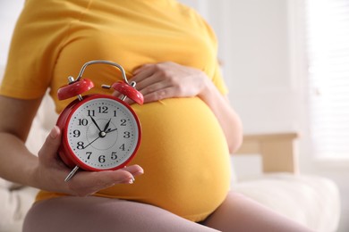 Young pregnant woman holding alarm clock near her belly at home, closeup. Time to give birth