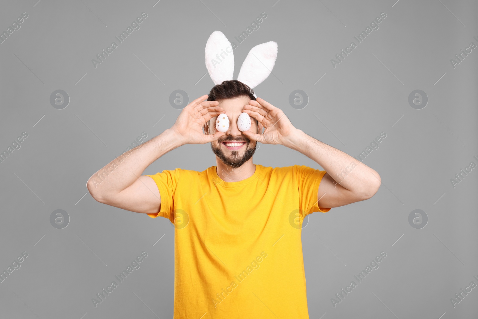 Photo of Happy man in bunny ears headband holding painted Easter eggs near his eyes on grey background