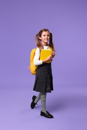 Smiling schoolgirl with backpack and book on violet background