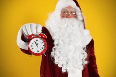 Photo of Santa Claus holding alarm clock on yellow background, focus on hand. Christmas countdown