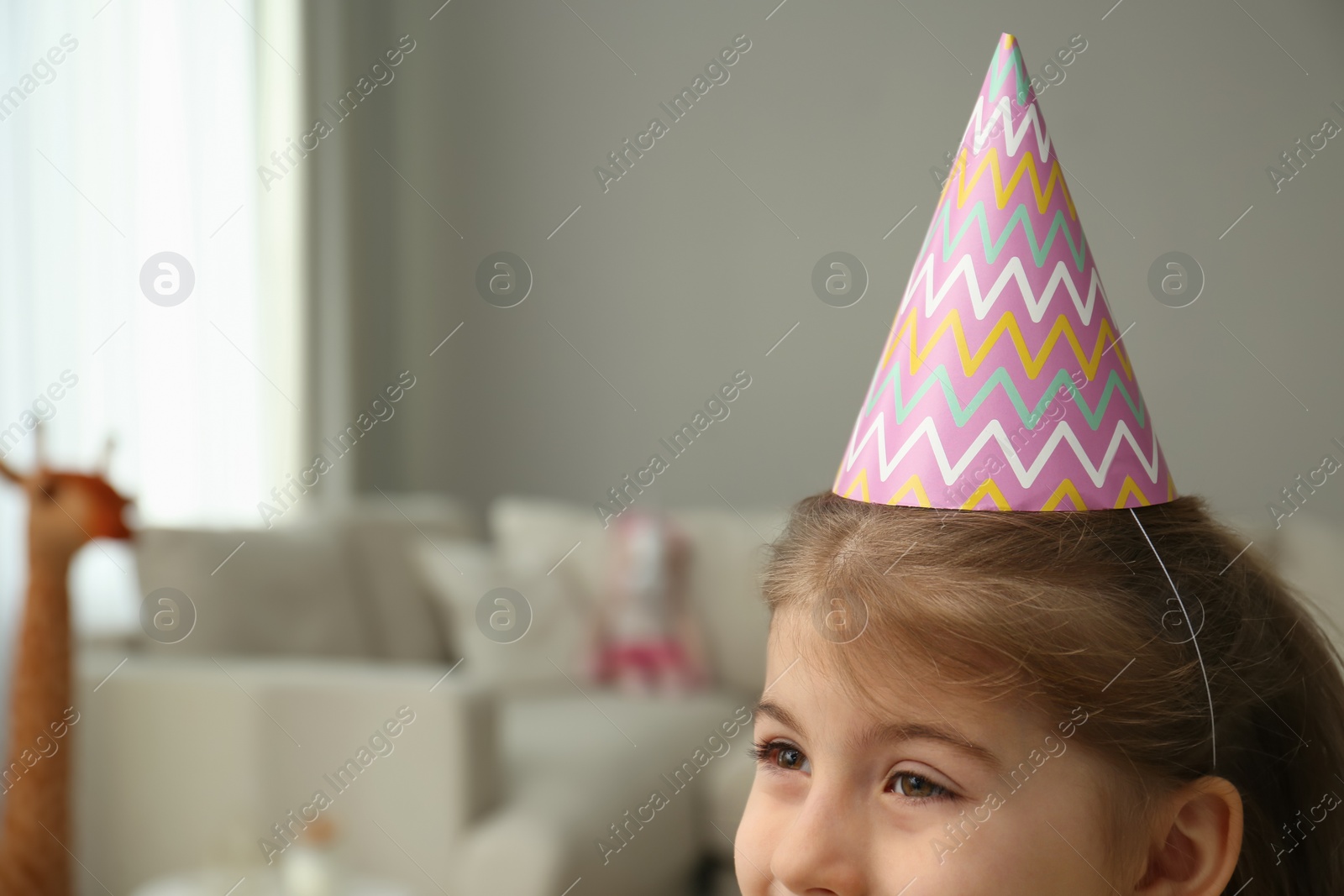 Photo of Cute little girl wearing party hat at home, space for text