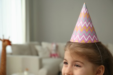 Cute little girl wearing party hat at home, space for text