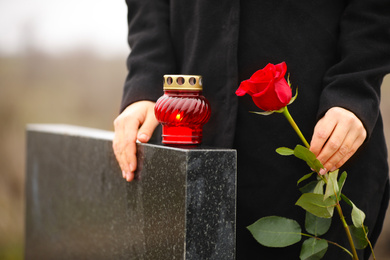 Photo of Woman holding red rose near black granite tombstone with candle outdoors, closeup. Funeral ceremony