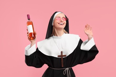 Photo of Happy woman in nun habit and sunglasses holding bottle of wine on pink background