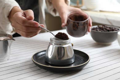 Photo of Woman putting ground coffee into moka pot at table in kitchen, closeup
