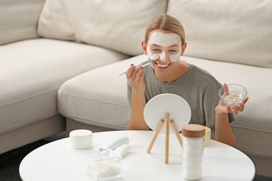 Young woman applying face mask in front of mirror at home, space for text. Spa treatments