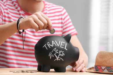 Photo of Man putting coin into piggy bank with word TRAVEL at table, closeup
