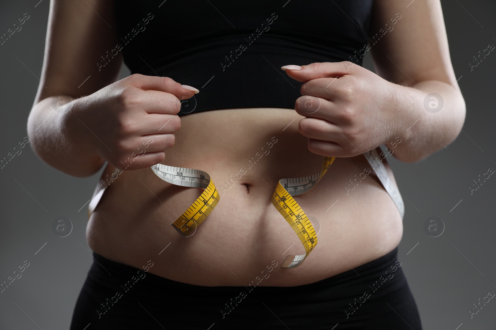 Photo of Woman measuring belly with tape on grey background, closeup. Overweight problem