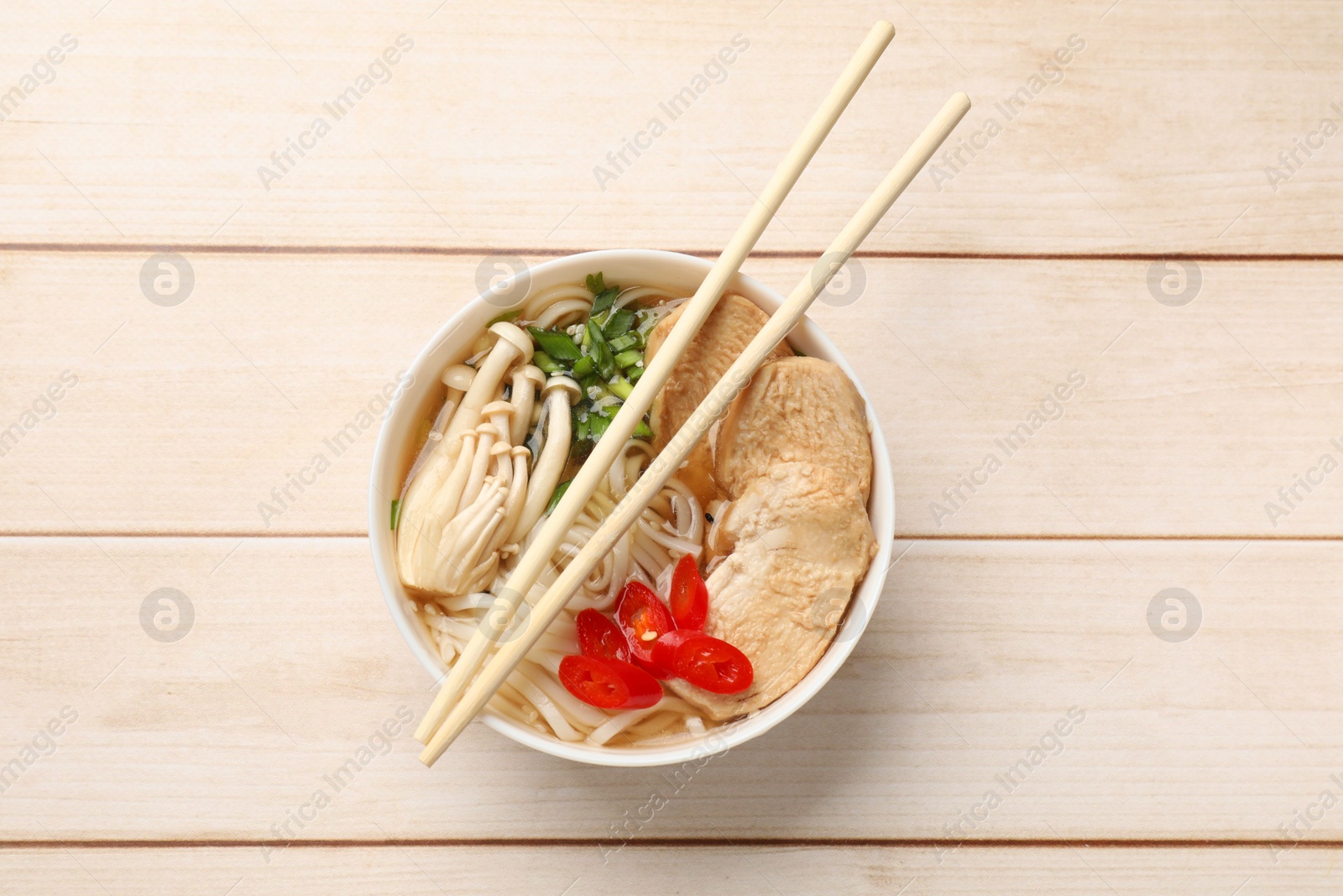 Photo of Delicious ramen with meat and mushrooms in bowl and chopsticks on white wooden table, top view. Noodle soup