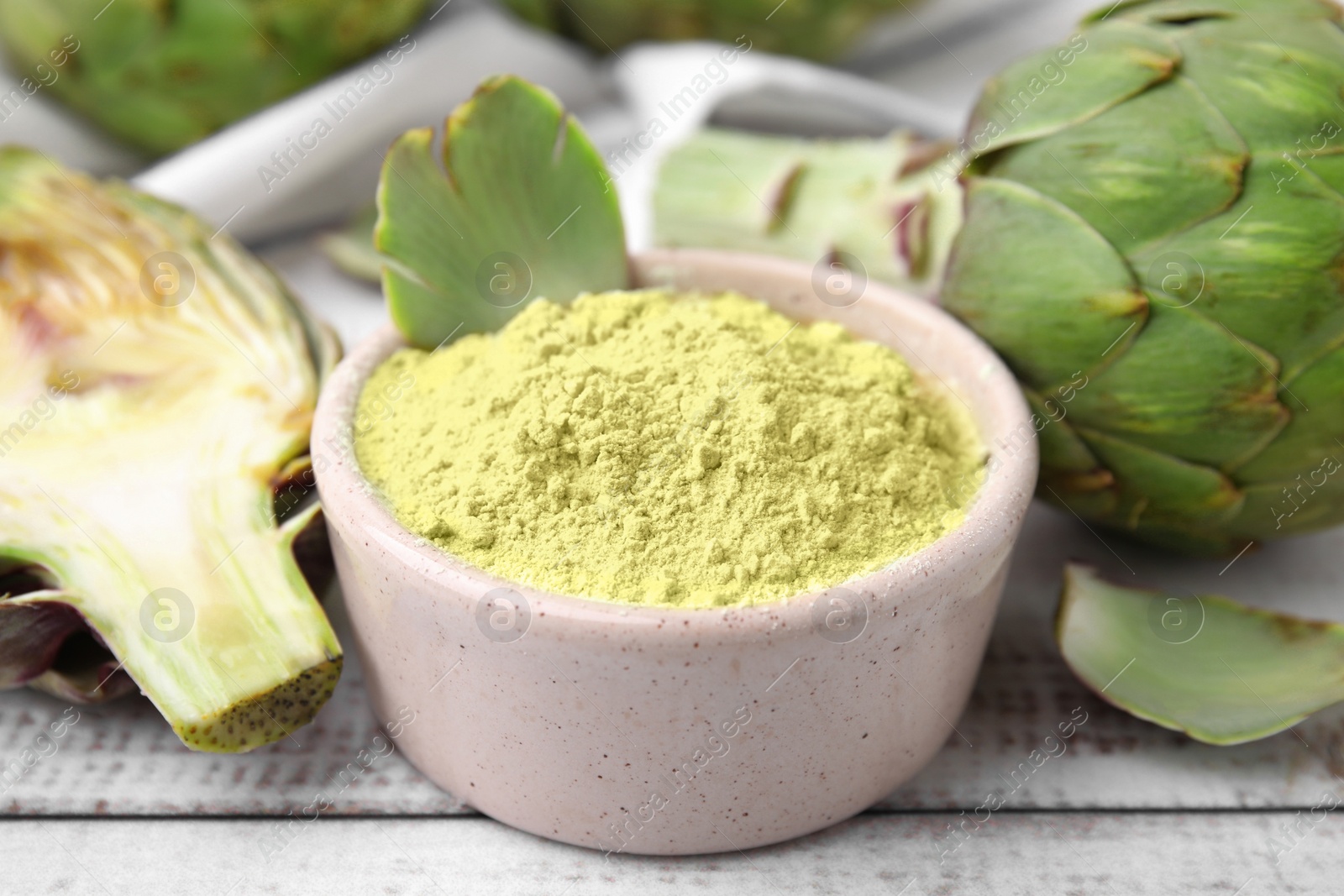 Photo of Bowl with powder and fresh artichokes on white wooden table, closeup