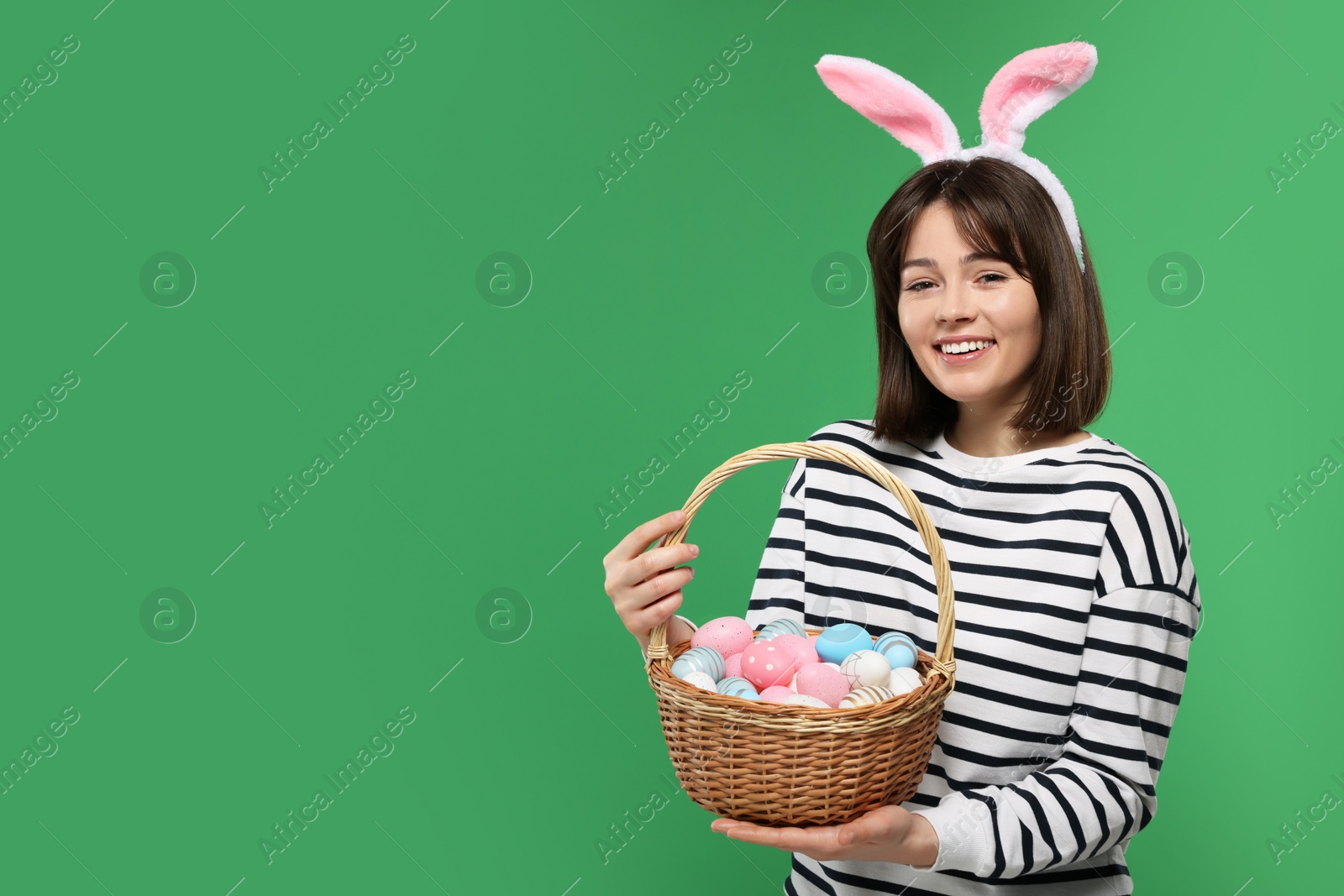 Photo of Easter celebration. Happy woman with bunny ears and wicker basket full of painted eggs on green background, space for text