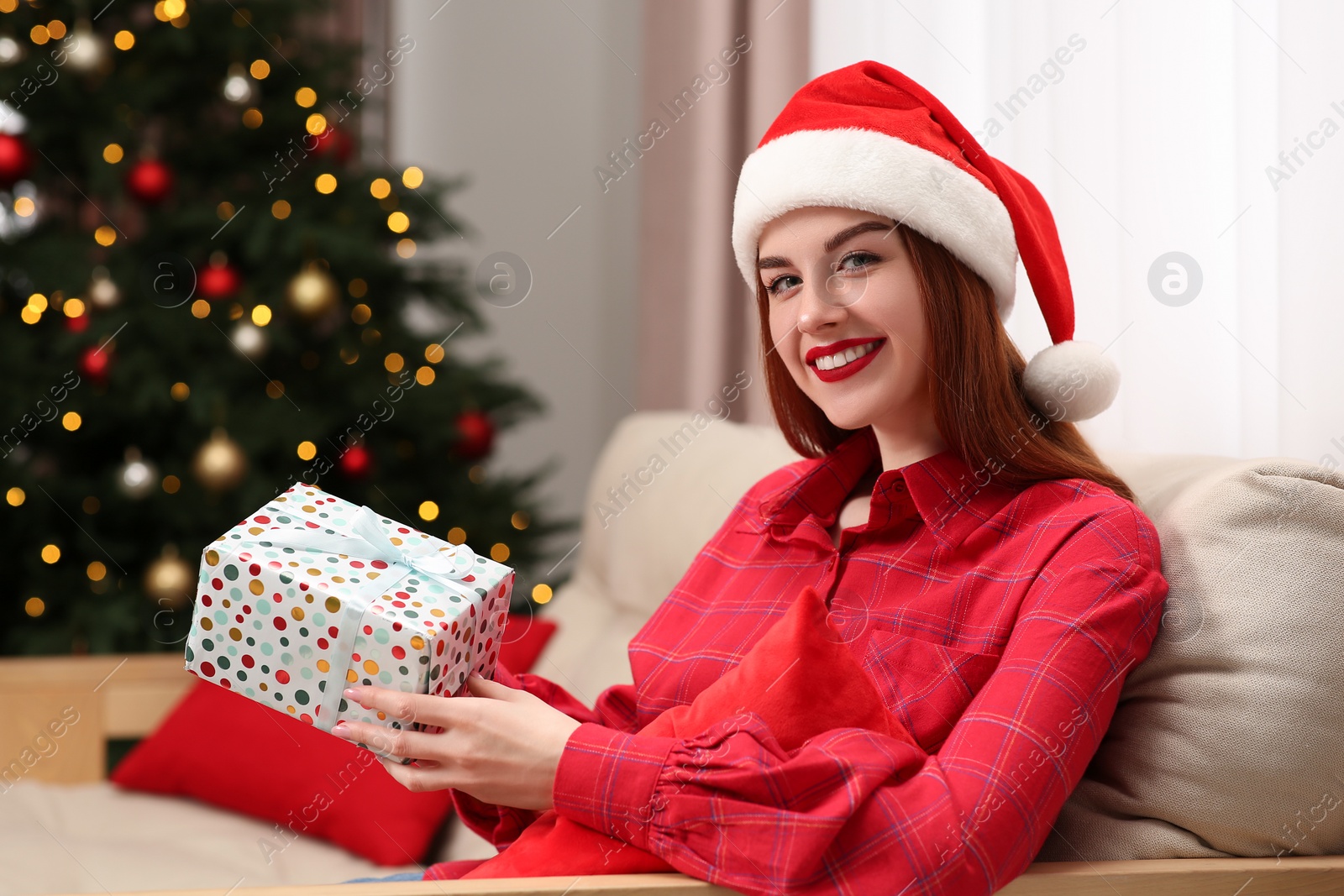 Photo of Beautiful young woman in Santa hat with gift box near Christmas tree at home
