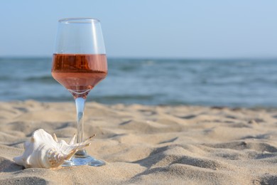 Glass of tasty rose wine and seashell on sand near sea, space for text
