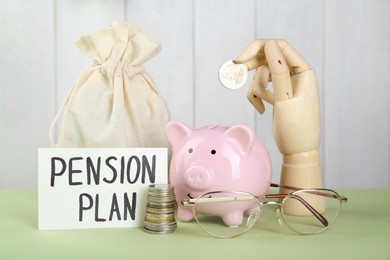 Card with phrase Pension Plan, wooden mannequin hand holding coin and piggy bank on light green table. Retirement concept