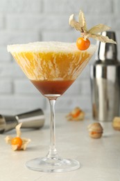 Photo of Delicious cocktail decorated with physalis fruit on light table near white brick wall