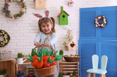 Photo of Adorable little girl with bunny ears and basket full of toy carrots in Easter photo zone