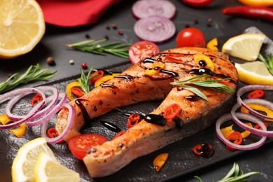 Photo of Slate plate with tasty salmon steak and vegetables on black table, closeup