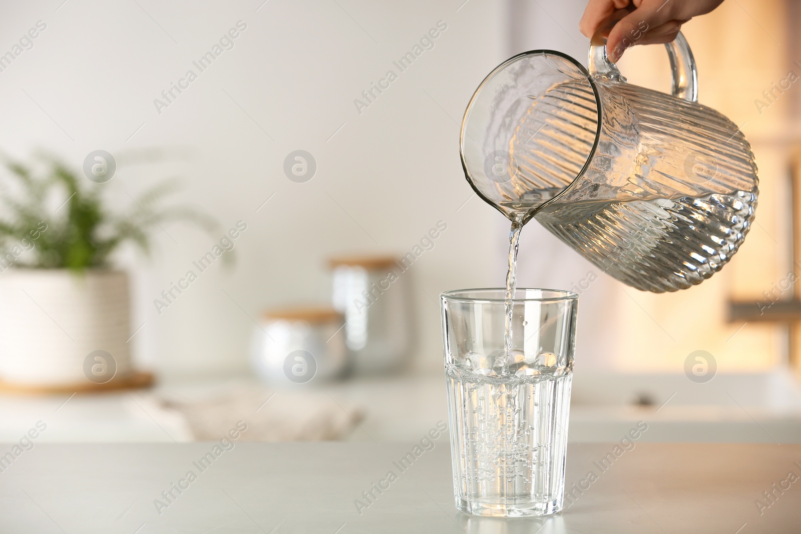 Photo of Woman pouring water from jug into glass at white table in kitchen, closeup. Space for text