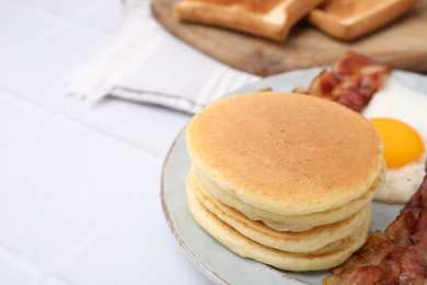 Photo of Plate with tasty pancakes, fried egg and bacon on white tiled table, closeup. Space for text