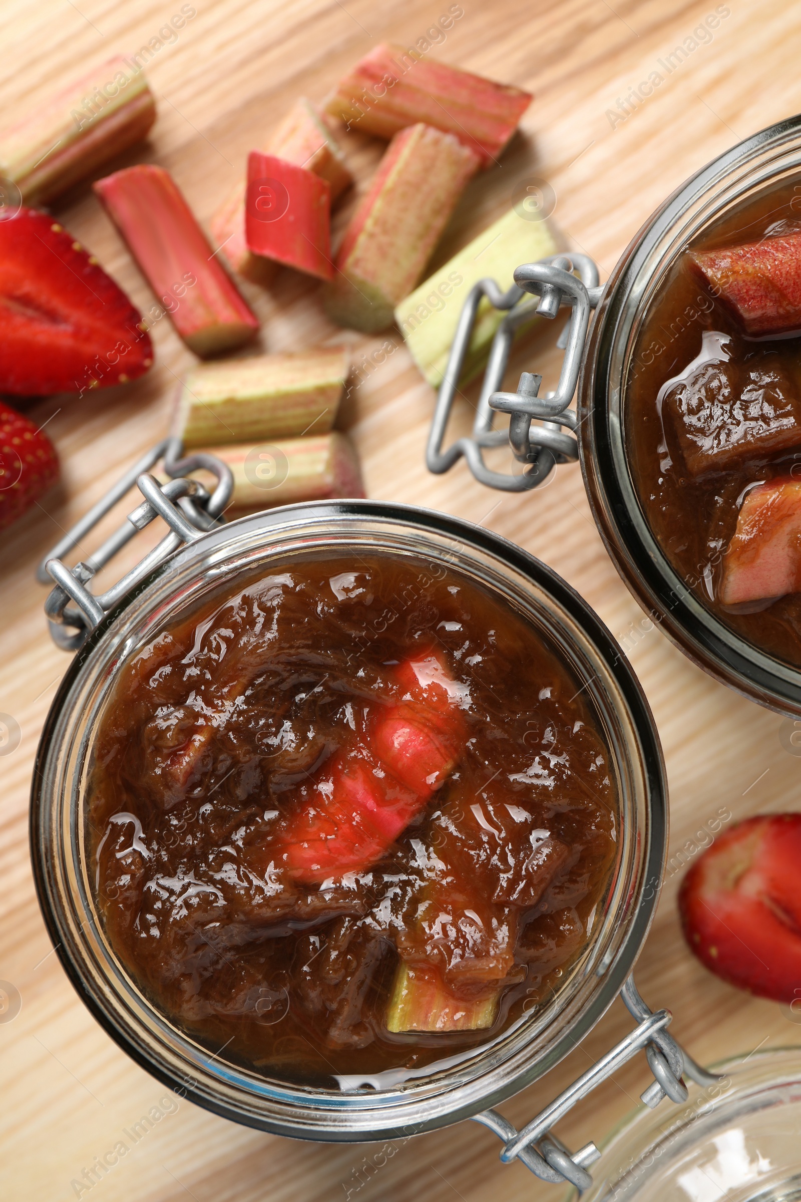 Photo of Jars of tasty rhubarb jam, strawberries and cut stems on wooden table, top view