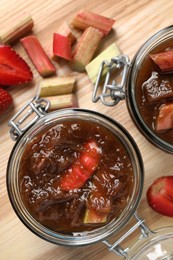 Jars of tasty rhubarb jam, strawberries and cut stems on wooden table, top view