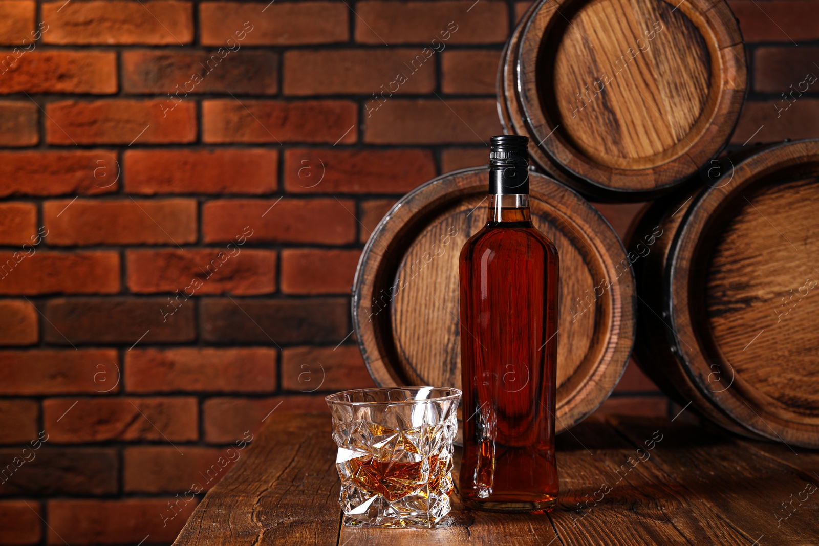 Photo of Glass and bottle of whiskey with wooden barrels on table against brick wall, space for text