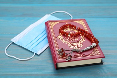 Muslim prayer beads, Quran and protective mask on light blue wooden table, closeup