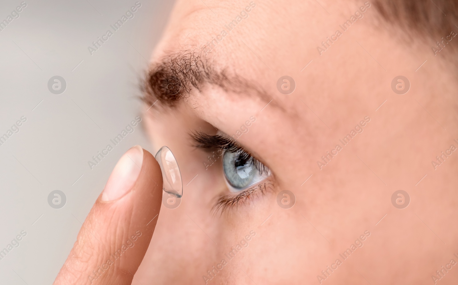 Photo of Young man putting contact lens in his eye on light background