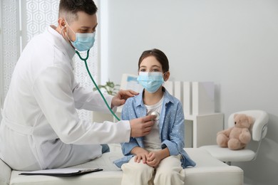 Photo of Pediatrician examining little girl in hospital. Doctor and patient wearing protective masks