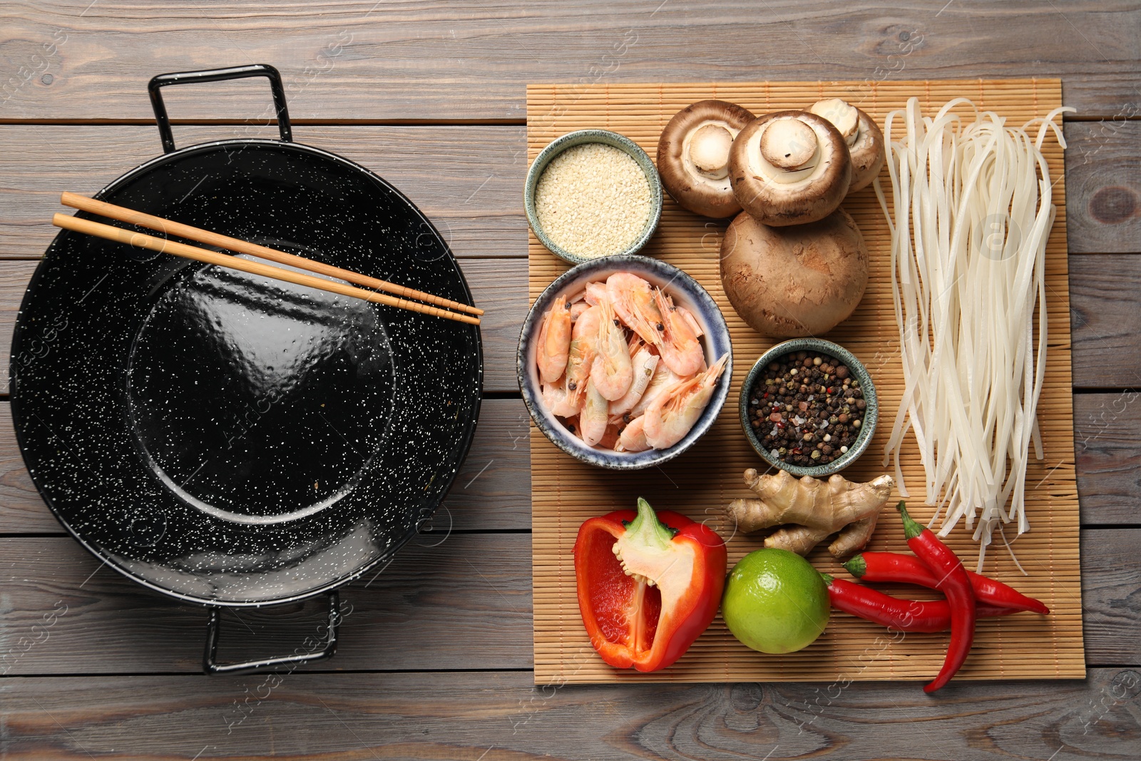 Photo of Black wok, chopsticks and bamboo mat with products on color wooden table, top view