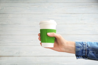 Woman holding takeaway paper coffee cup with cardboard sleeve on light background