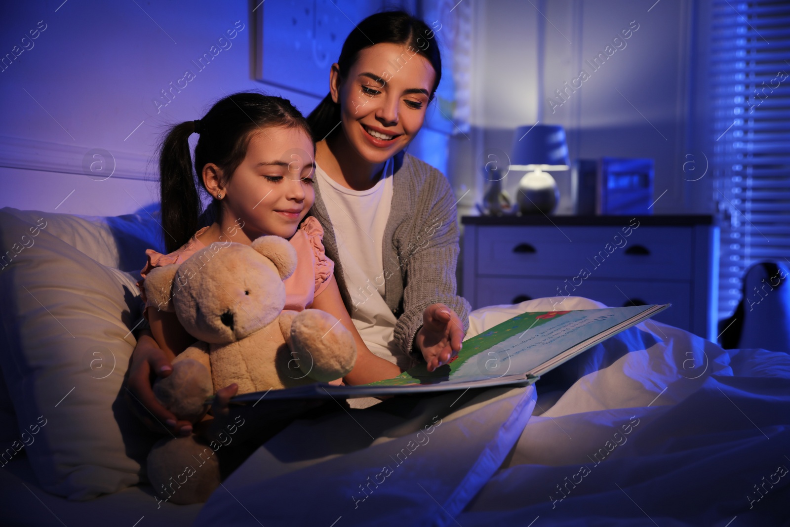 Photo of Mother with little daughter reading fairy tale in dark bedroom