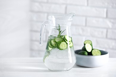 Refreshing cucumber water with rosemary in jug and vegetables on white table against brick wall. Space for text