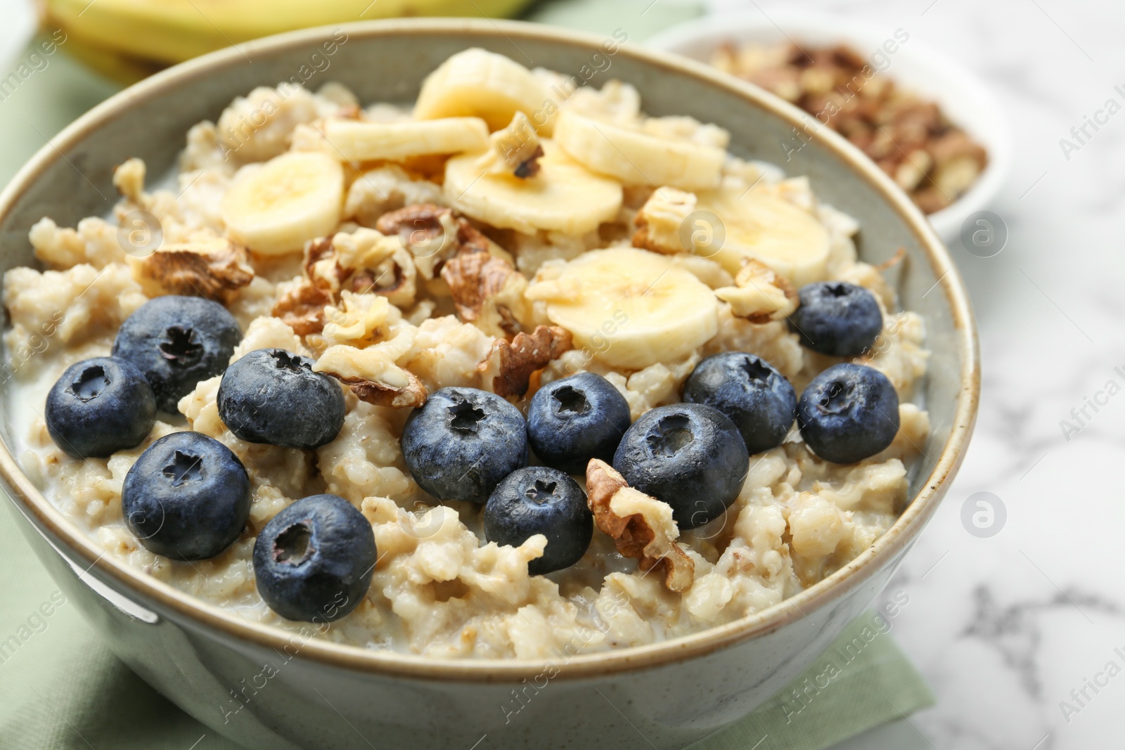 Photo of Tasty oatmeal with banana, blueberries, walnuts and milk served in bowl on white marble table, closeup