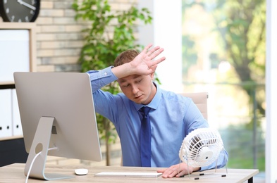 Photo of Young man suffering from heat in front of small fan at workplace