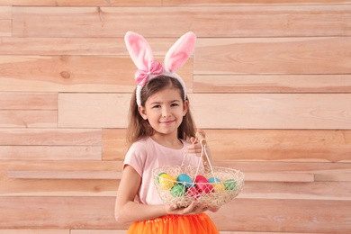 Photo of Little girl in bunny ears headband holding basket with Easter eggs against wooden background, space for text