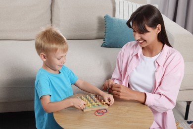 Photo of Motor skills development. Happy mother helping her son to play with geoboard and rubber bands at coffee table in room