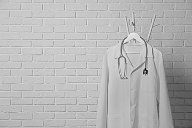 Photo of Medical uniform and stethoscope hanging on rack near white brick wall. Space for text