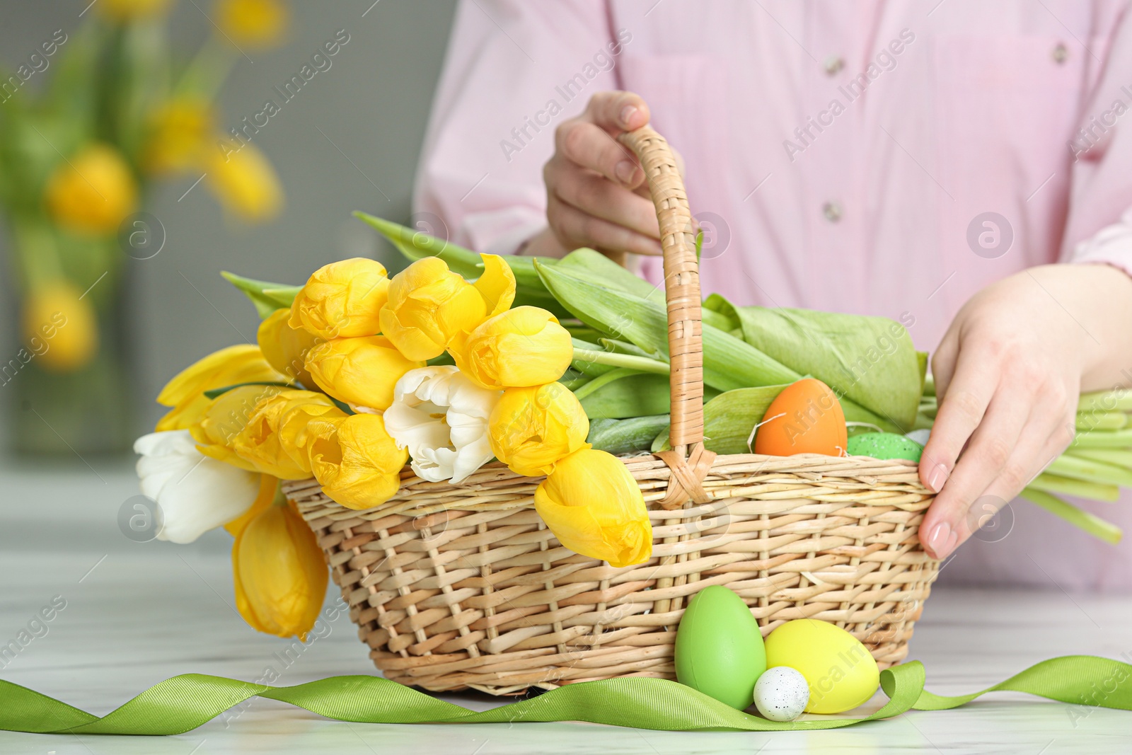 Photo of Woman taking Easter basket with bouquet of tulips and eggs at white marble table indoors, closeup