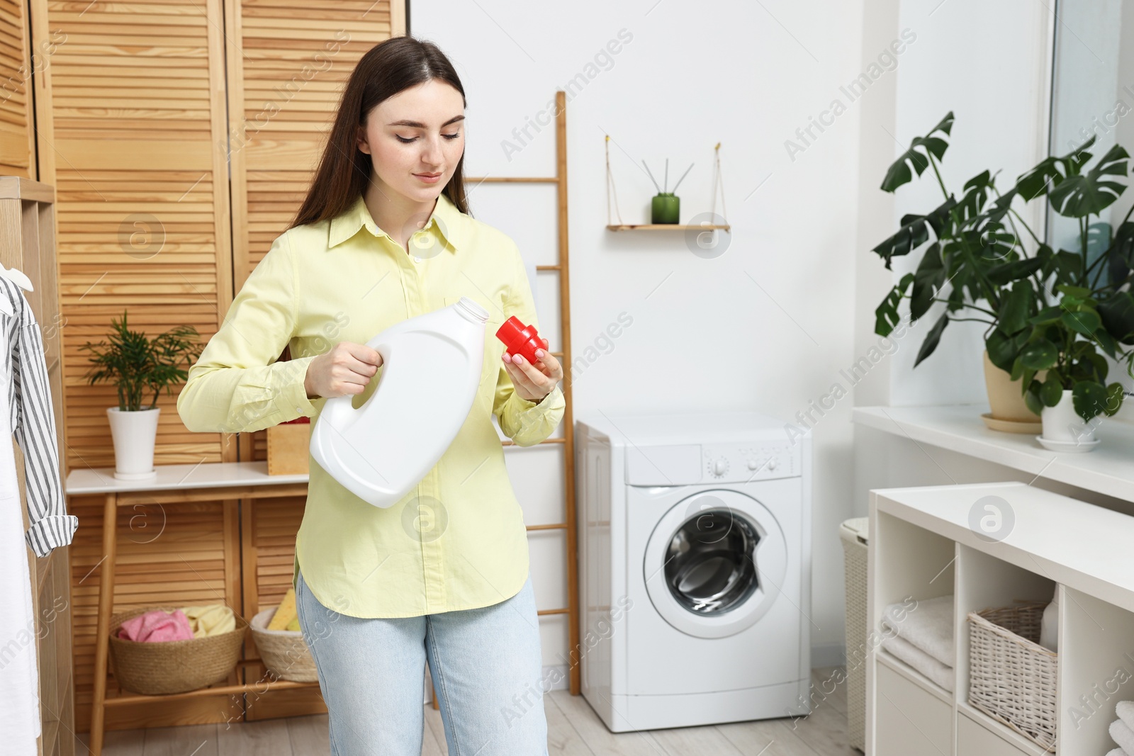 Photo of Beautiful young woman pouring detergent into cap in laundry room